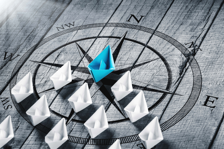 White paper boats arranged in triangular formation on wooden compass, with blue boat in the lead