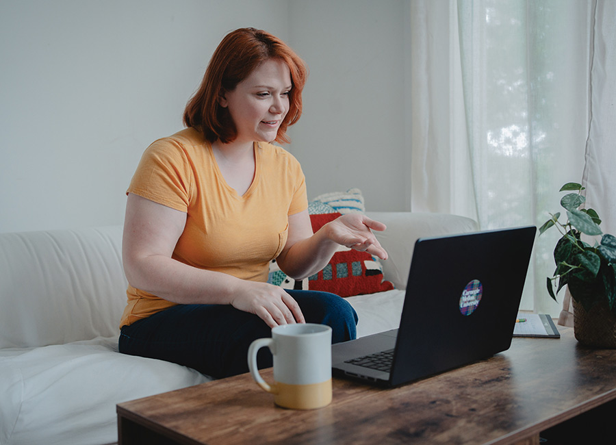 female student with red hair is sitting on couch, pointing at a laptop computer, and working on an assignment for her masters degree in innovation online