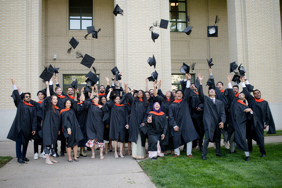 students stand in front of a building in graduation attire throwing their caps into the air