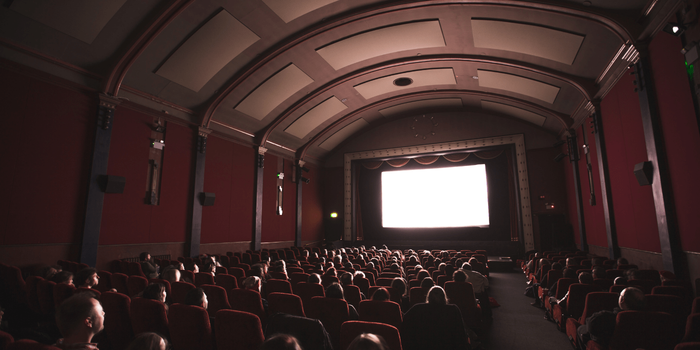 A dark movie theater with people sitting in seats watching a lit-up screen