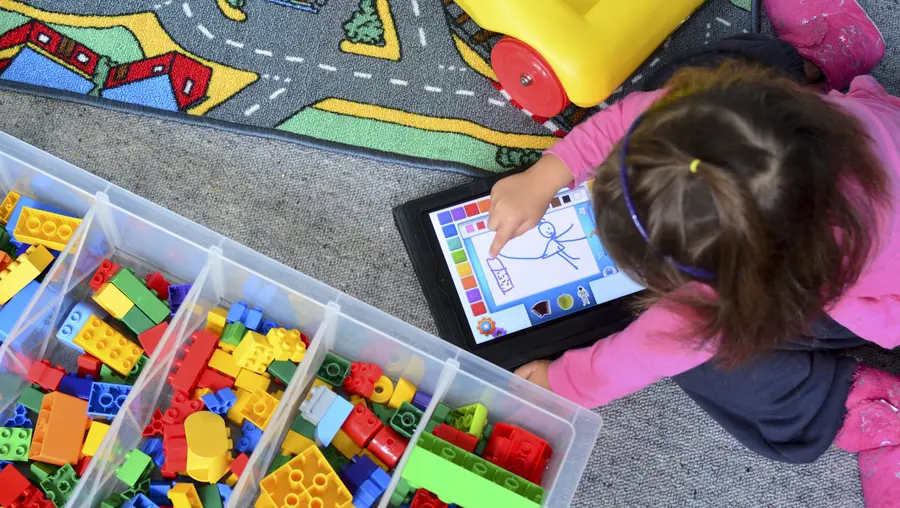 a student sits on the floor and plays with a drawing application on a tablet next to bins of colorful blocks