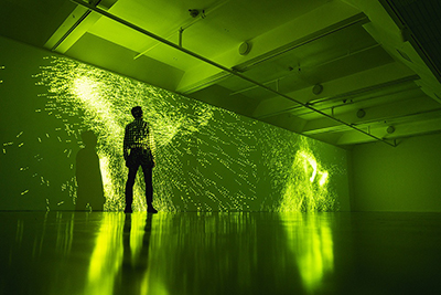 Image of a person standing in front of a long wall with neon green light projections of a splattered shape covering the wall and person in a dark room.