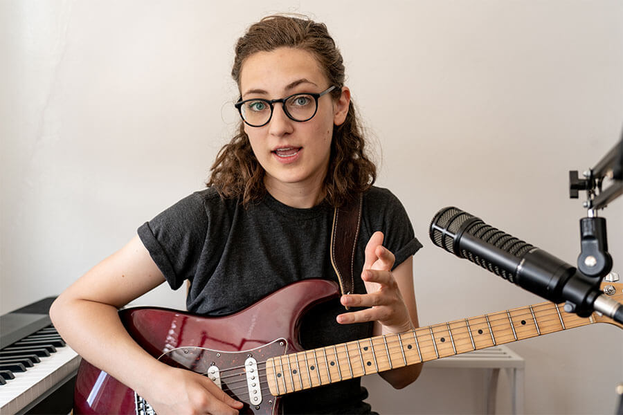 Image of BriTE music instructor Heather DiCicco holding guitar next to piano keyboard while teaching a Zoom music class