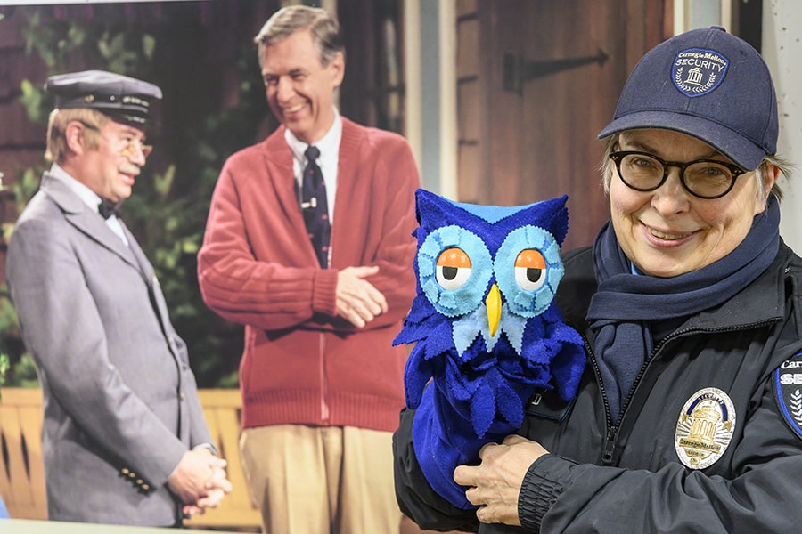 image of Kathy Borland with X the Owl standing in front of a poster of Fred Rogers