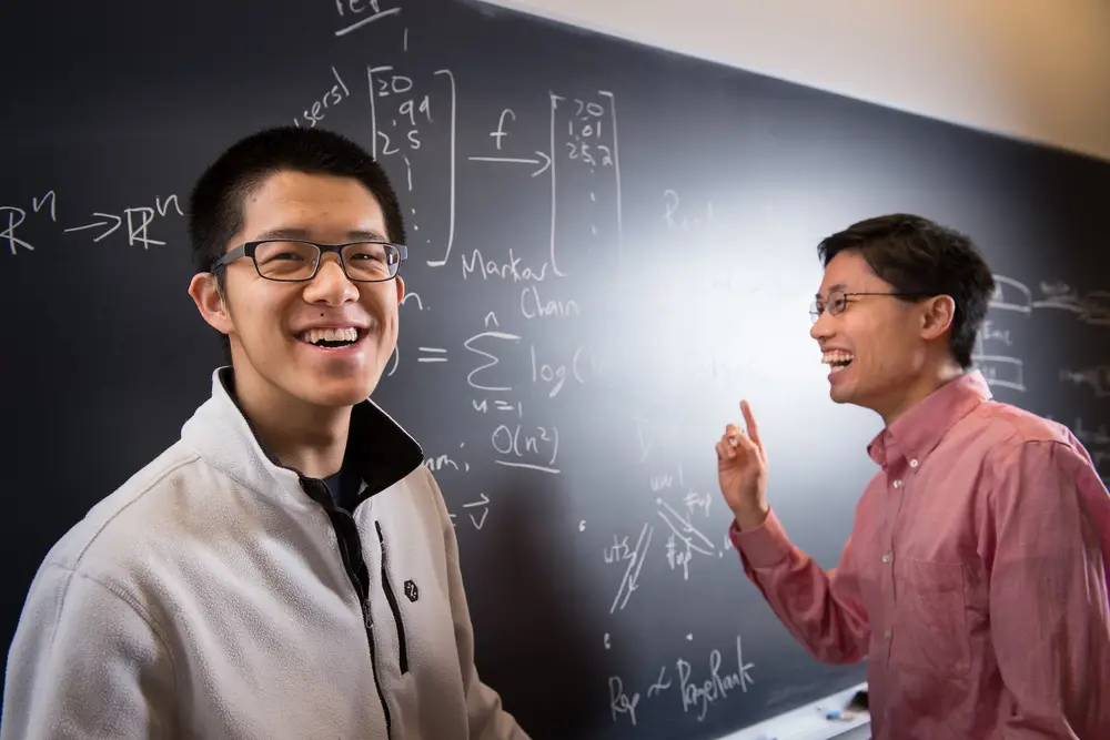 Student smiling as professor Po-Shen Lo smiles and points to the whiteboard full of math equations.