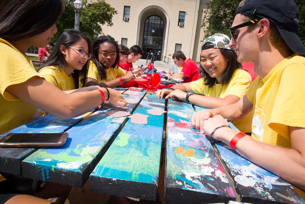 Students sitting at a picnic table outside, playing cards.