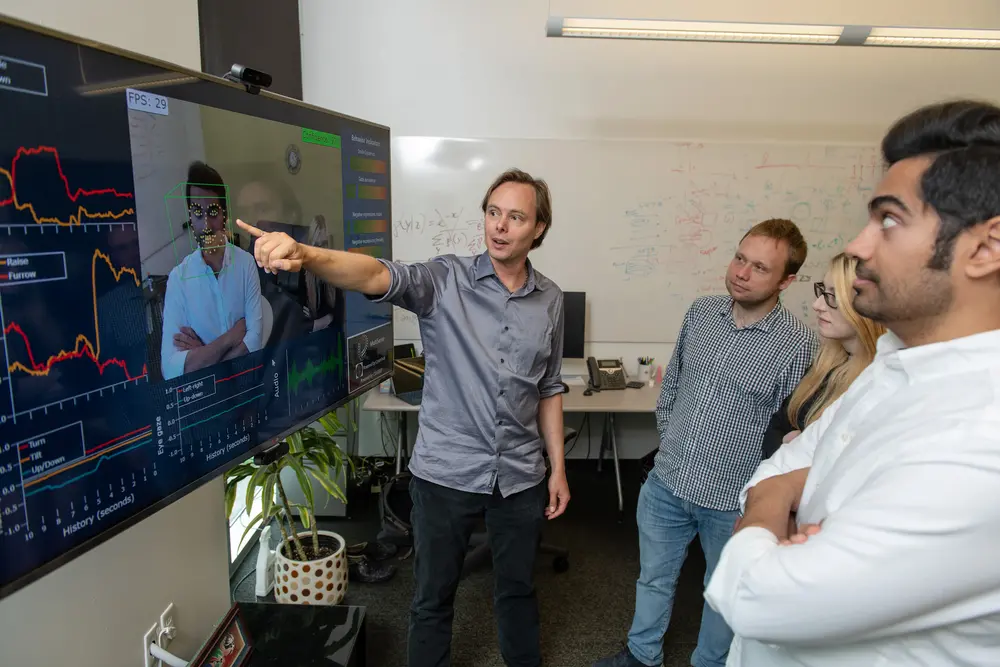 Students watch as professor LP Morency points to a TV that is capturing data from their facial expressions.