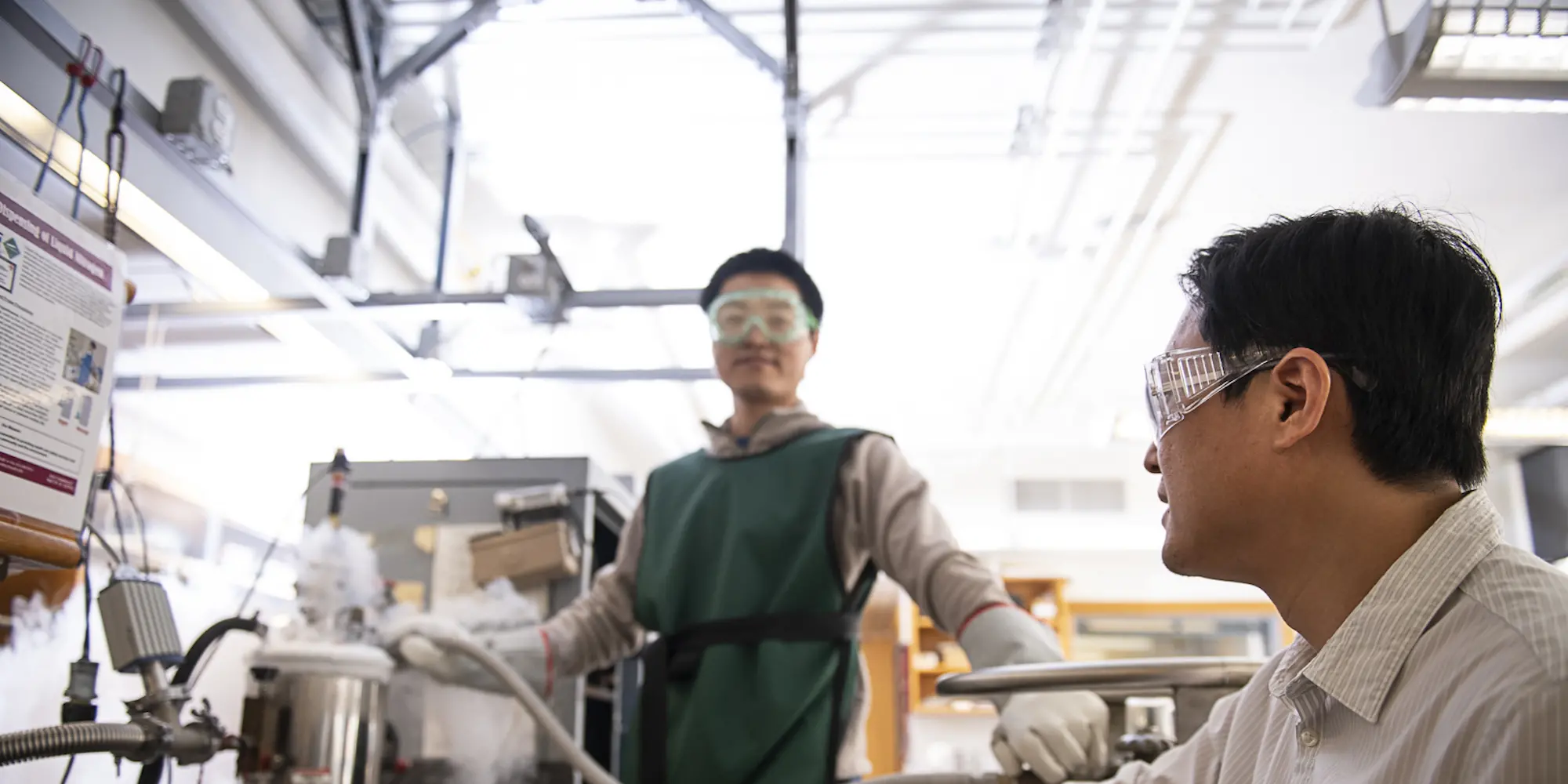 Student in a lab wearing protective lab gear, working with a cylinder of gas as professor Yisong Guo watches.