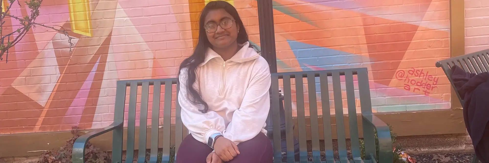 A photo of Samhita sitting on a bench in front of a mural in Shadyside.