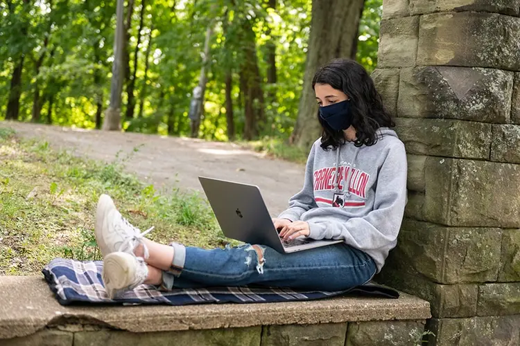 photo of student leaning against a stone wall with laptop and mask