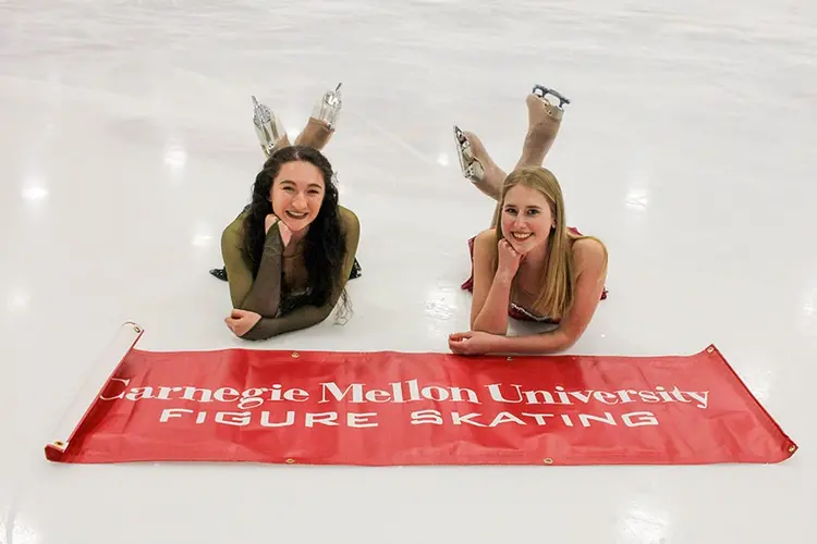Two CMU figure skaters laying on the ice behind a CMU Figure Skating red banner