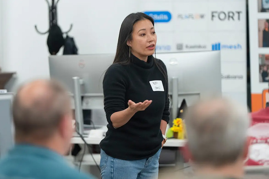 A woman in a black turtleneck sweater speaking and gesturing to a group during a presentation.