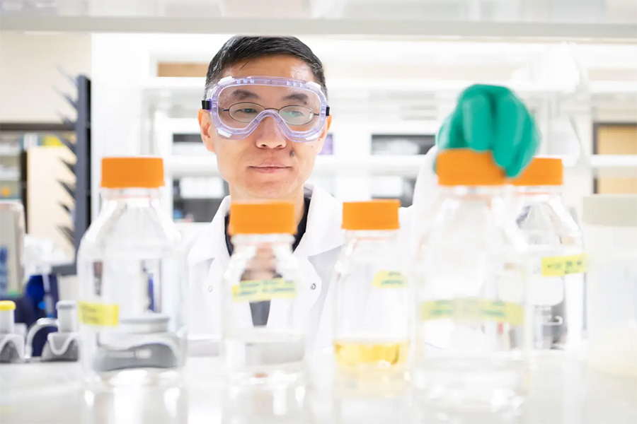 A male researcher wearing protective goggles and a white lab coat reaches with a green-gloved hand toward orange-capped glass bottles stored on a laboratory shelf.