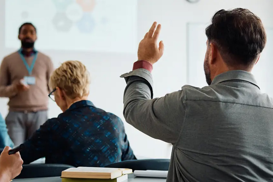 View from behind an audience member raising his hand to ask a question while a presenter stands near a projection screen at the front of a brightly lit room.