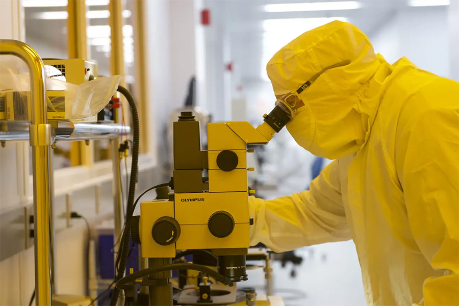A researcher wearing a full yellow cleanroom suit and safety goggles looks into a high-powered microscope in a nanotechnology laboratory.