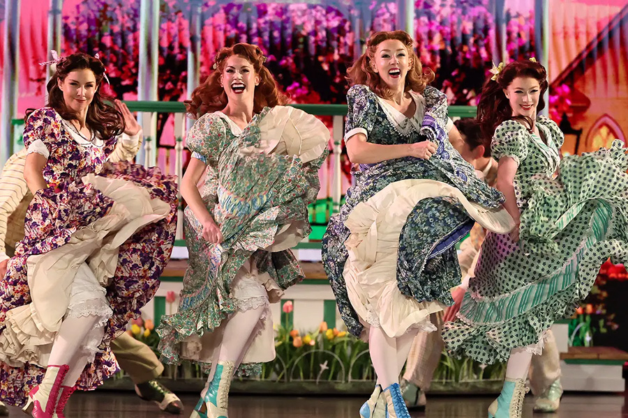 Four women performing a lively high-kicking dance in colorful vintage floral dresses on a brightly decorated stage.