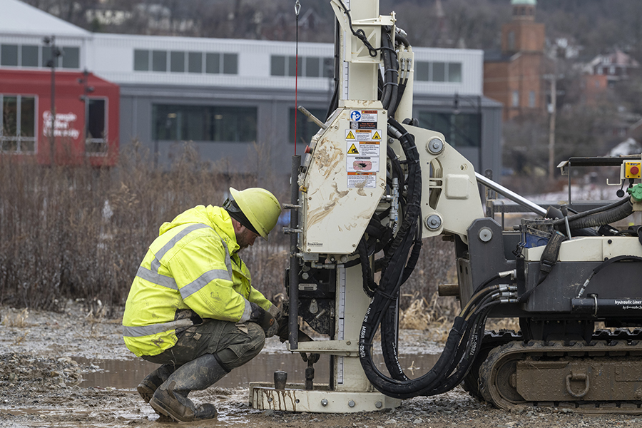 A man in a yellow vest and hardhat kneels in front of a piece of machinery that is digging deep in the soil.