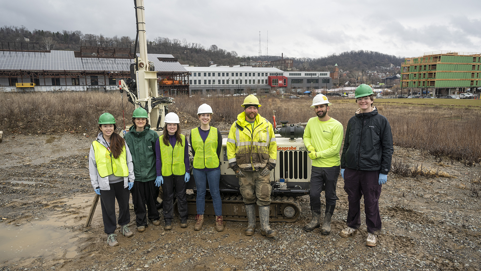 Seven people wearing hard hats and safety vests stand in front of a digging machine in an empty lot of dirt.