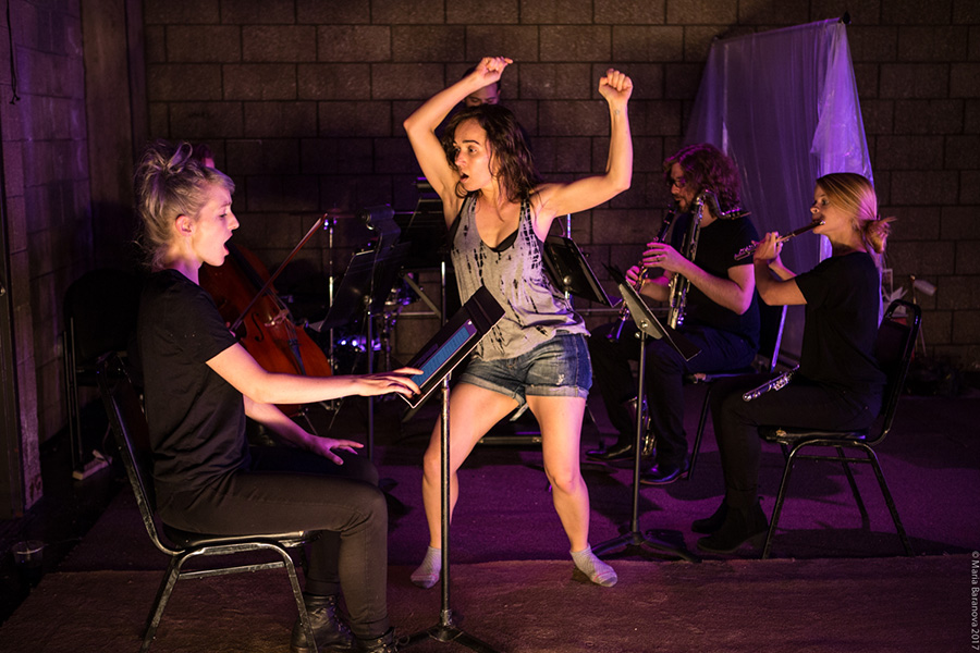 A woman dances as another woman sings while looking at music on a music stand. In the background musicians play in the dark