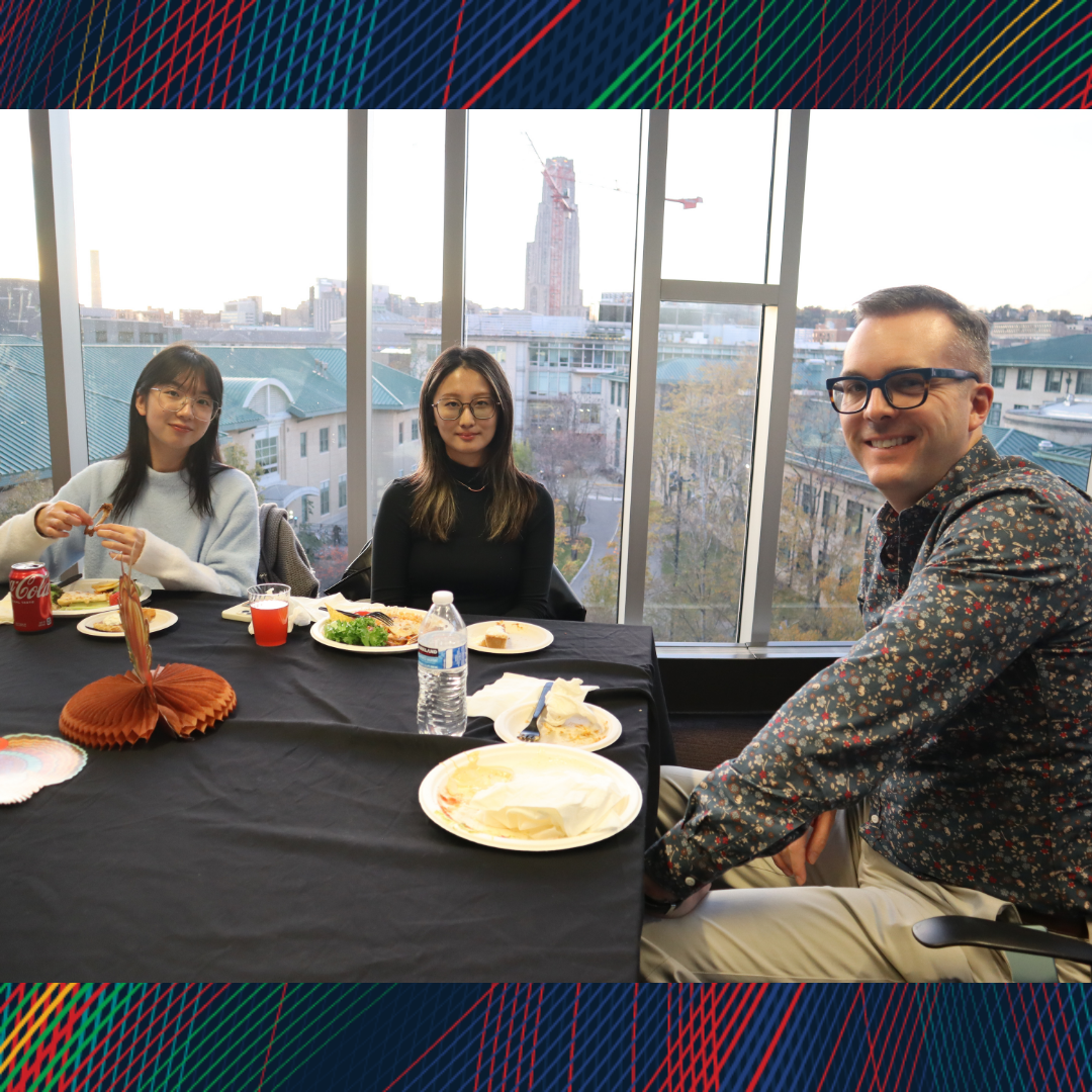 Two students and a professor smiling at the camera during dinner with the Cathedral of Learning in the background.