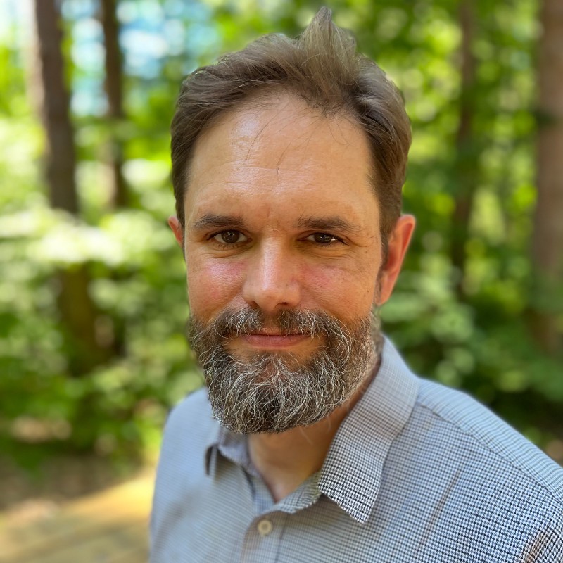 A headshot of a light haired man with a beard standing in front of a green background