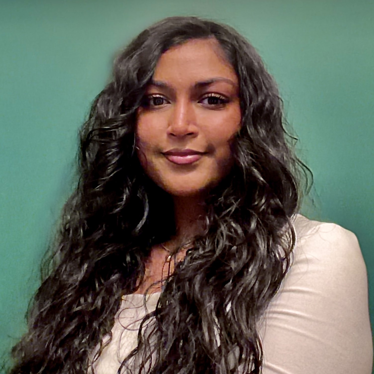 This image contains a woman with dark, wavy hair, wearing a beige shirt, standing in front of a green background