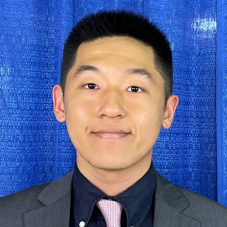 Headshot of a man with dark hair, wearing a suit and tie against a blue background