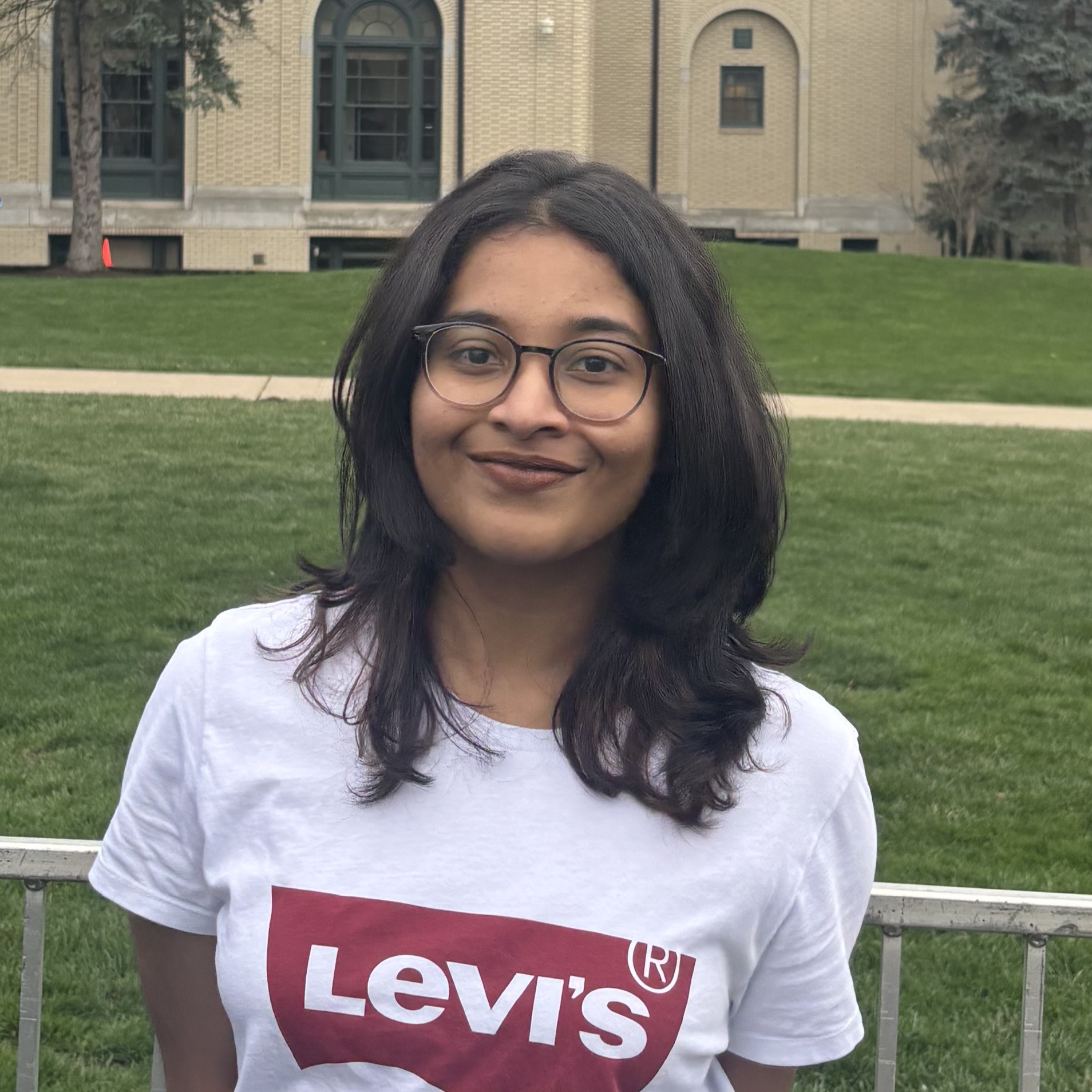 A dark haired woman wearing glasses and a white t-shirt with a red Levi's logo stands in front of an academic building