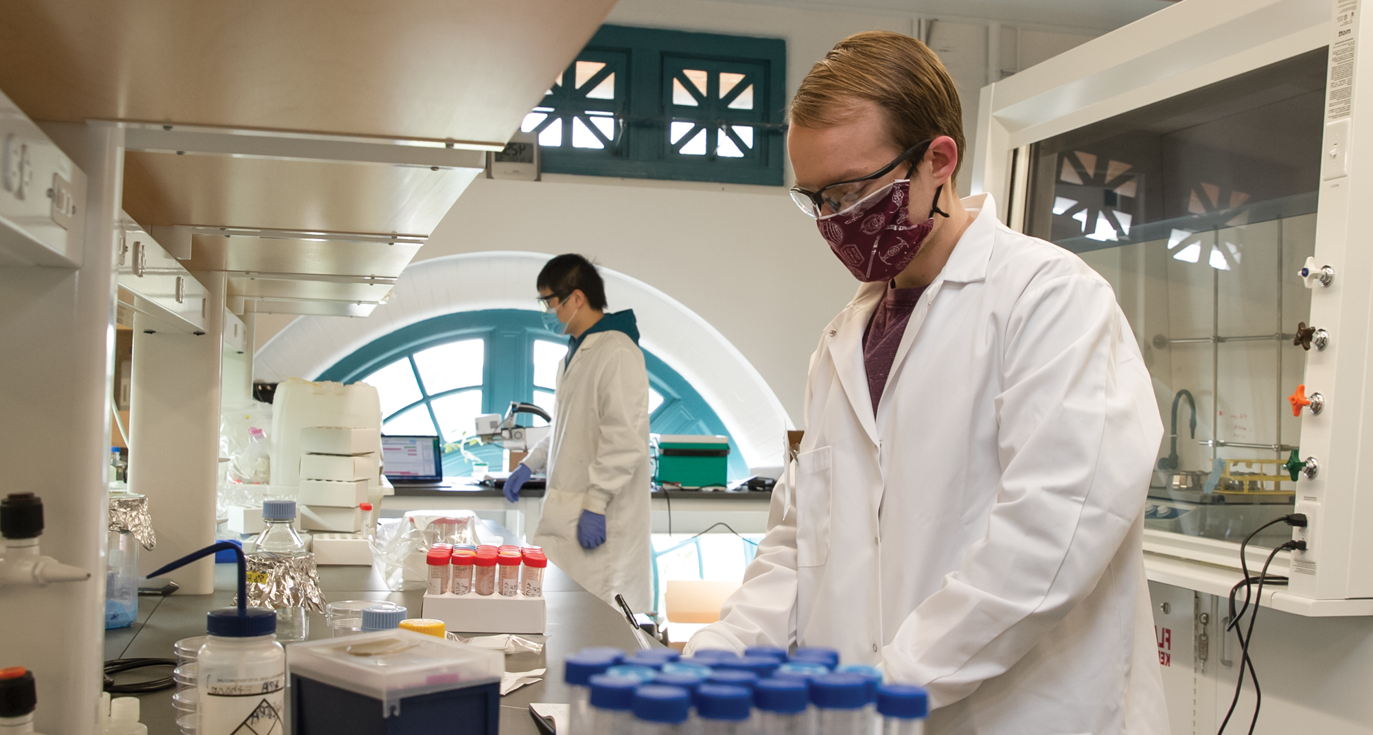 Two students are at a lab bench. In the foreground, Garrett Bland writes in his lab notebook and Yilin Zhang is in the background conducting an experiment
