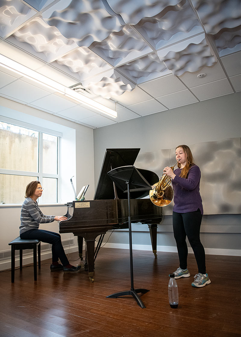 Photo of a student playing french horn for a professor, accompanying on a piano.