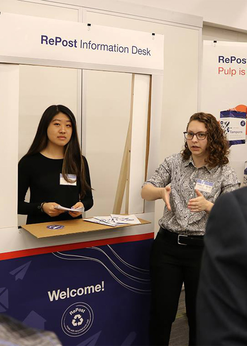 Two students presenting inside a cardboard replica of a postal service window.