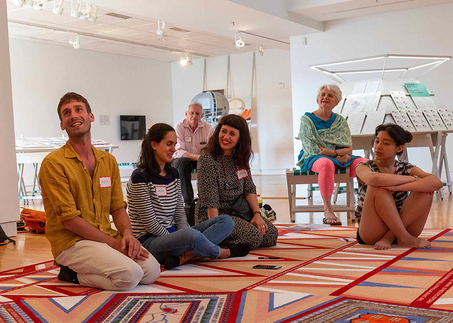 Students gathered around a welcoming rug, not wearing shoes. Two professors sit nearby. All are engaged, responding to something just off frame.