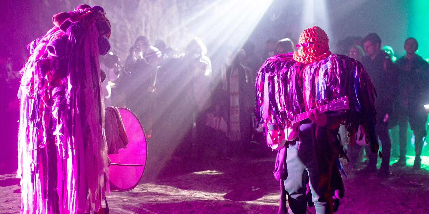 Photo of band wearing outfits with long strips of fabric dangling from them, bathed in magenta light.
