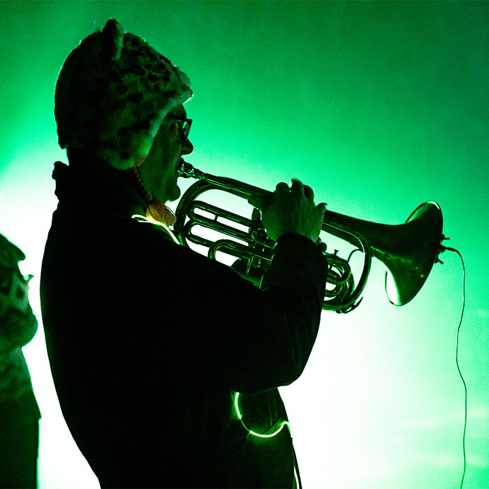 Man playing brass instrument and wearing animal hat, bathed in green light.