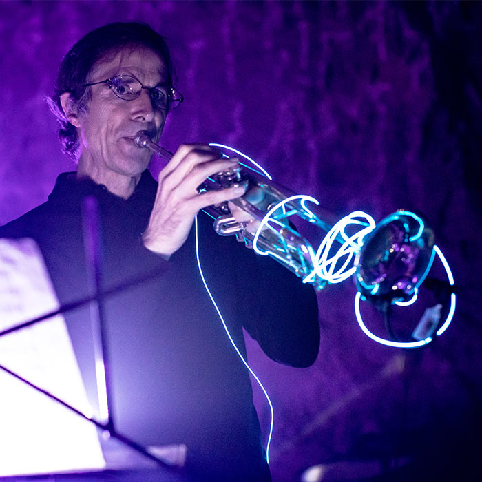 Man playing brass instrument covered in light blue rope lights with purple light in the background.