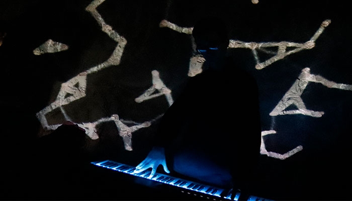 Man in dark lighting, playing a keyboard that glows blue, with white shapes projected on him and the limestone wall in the background.