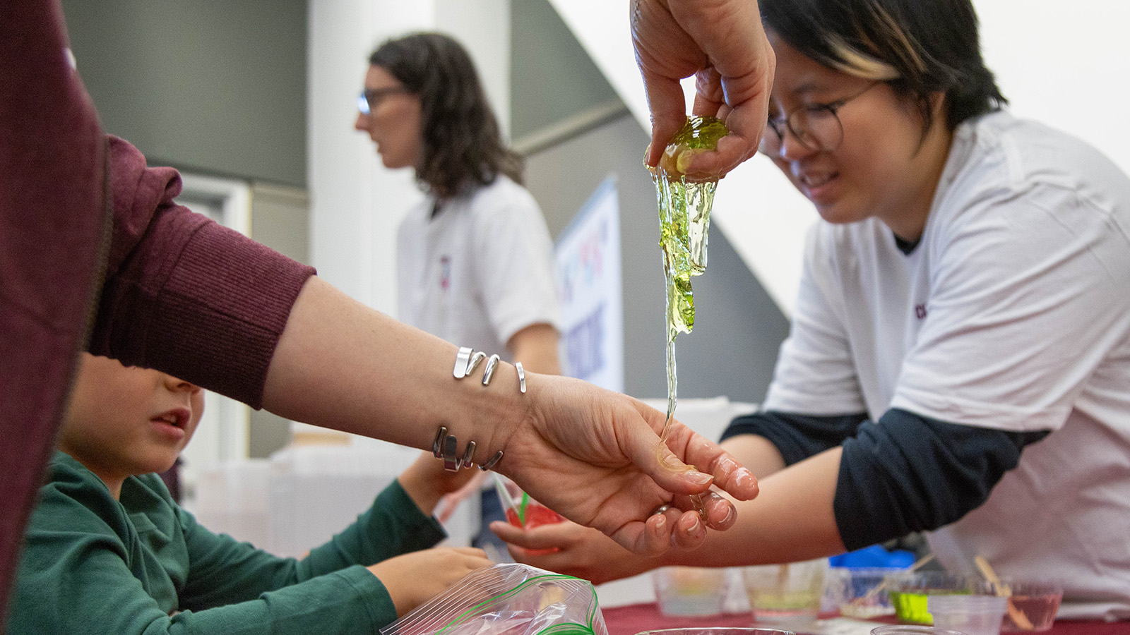 In the foreground, a person dangles a piece of slime. In the background, a graduate student places slime in a young person's hand