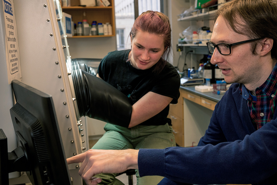 Image of a female student using a hood with gloves while a doctoral male student watches enzymes on a monitor
