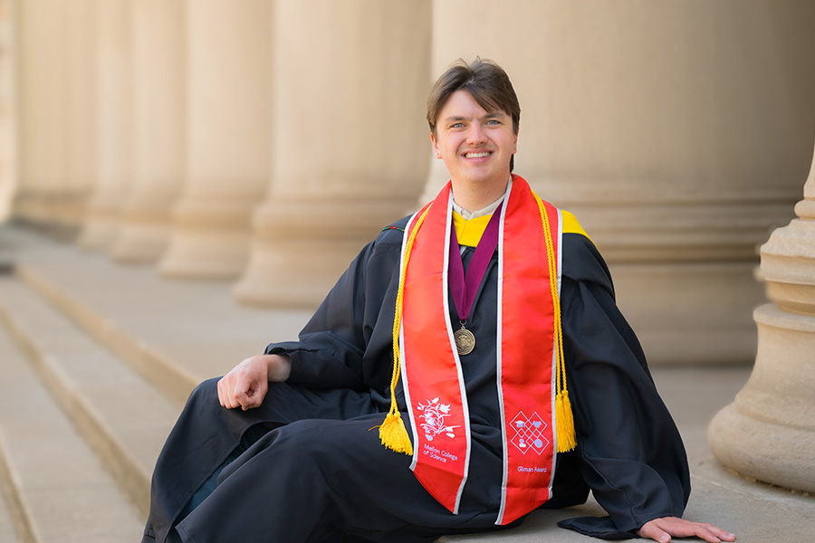 Photo of a male student in a Commencement gown in front of building columns