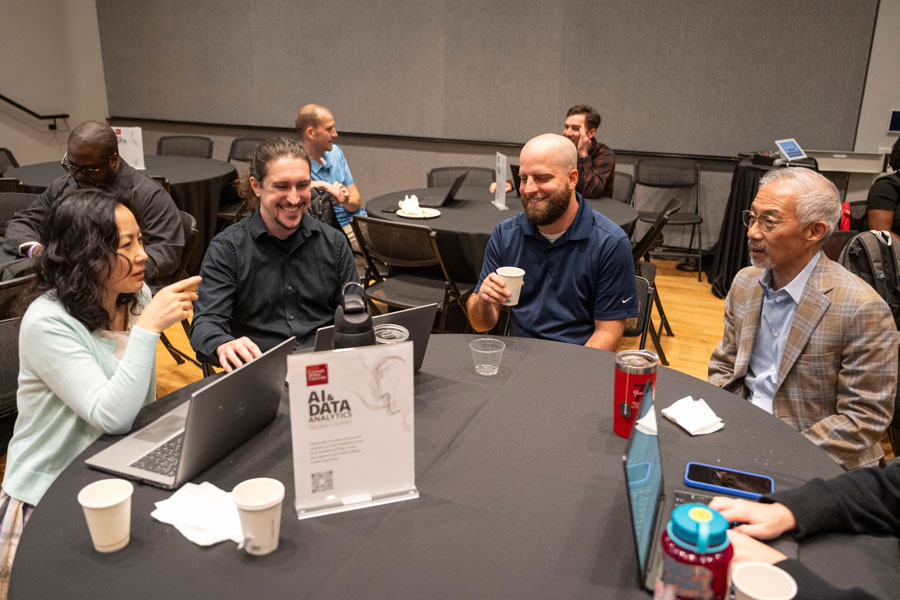 A group of diverse professionals converses and works on laptops at a round table with event materials.