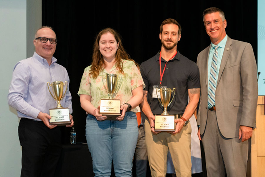 Three award winners smile on stage as they hold their first-place trophies.