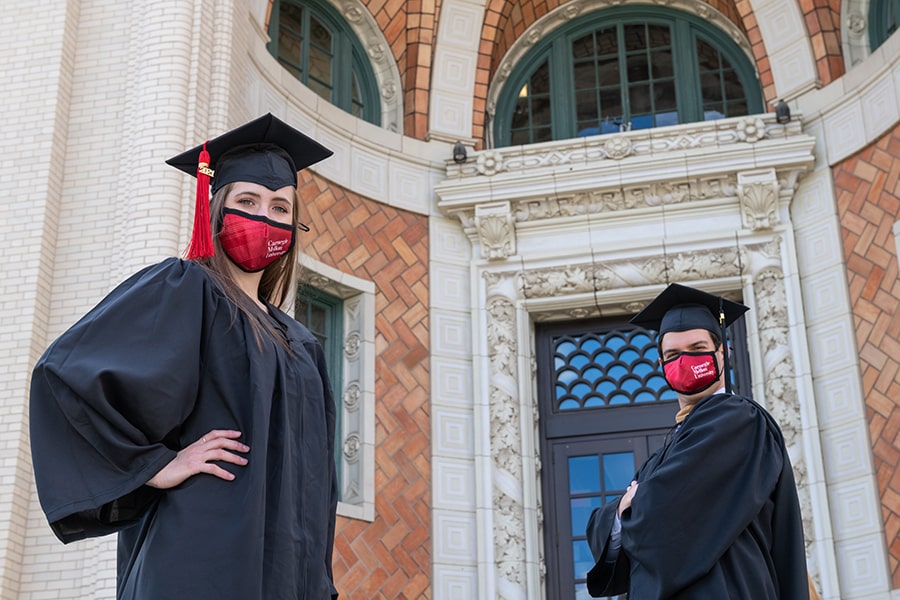 Grads on campus in caps and gowns, wearing facial coverings.
