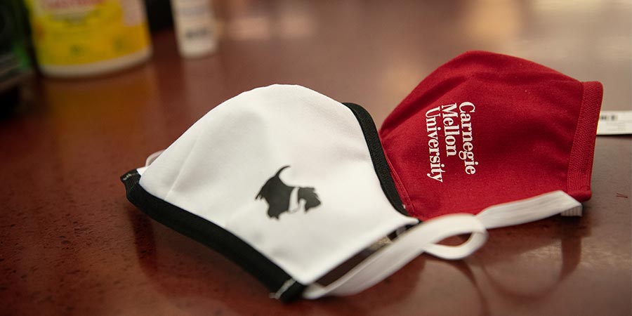 A white face mask with the CMU mascot, Scotty, and a red face mask with the white CMU wordmark on a wooden desk.