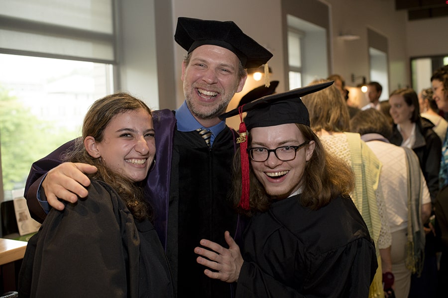 Professor Christopher Warren poses with two students during the English Department diploma ceremony. They are dressed in robes and smile excitedly.