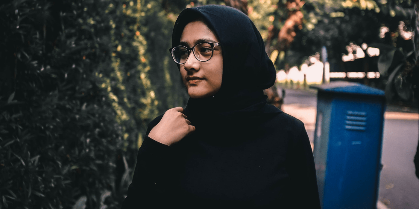 A young woman in glasses wearing a hijab looks to the side