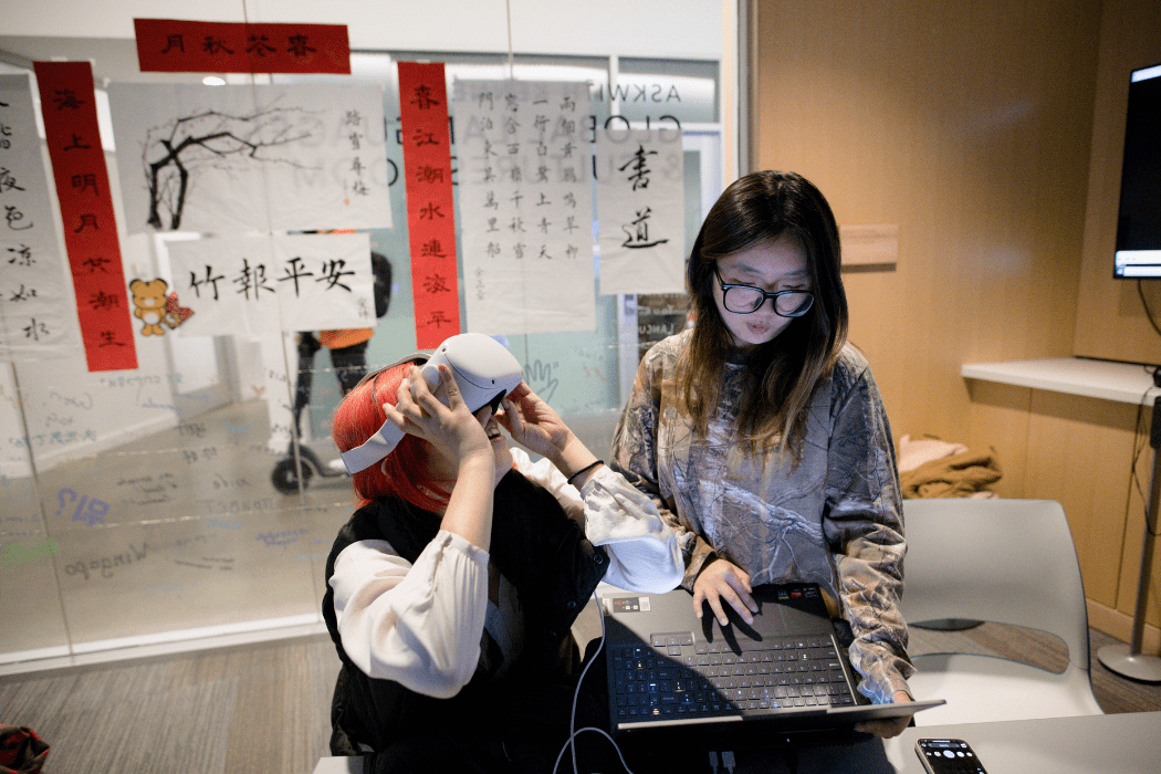 A person is seated wearing a VR headset. A second person stands next to them using a laptop.