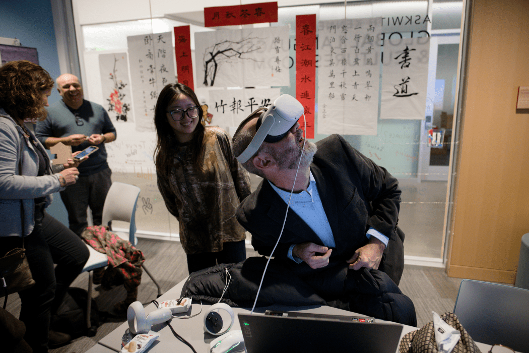 A man wears a VR headset and is looking upward. Three people in the background observe.