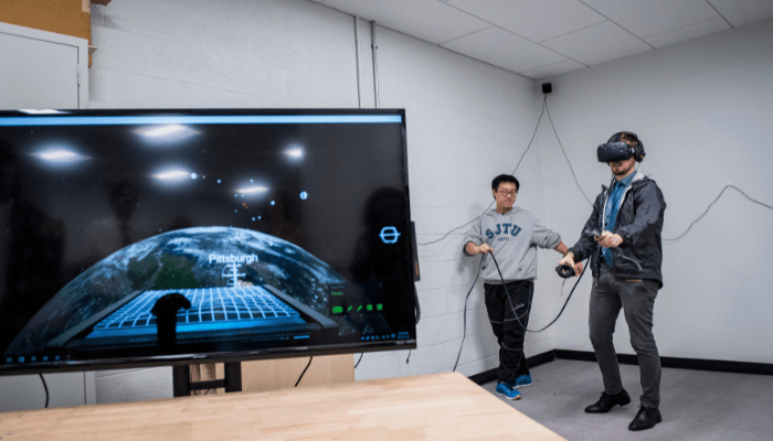 Two students stand in a room, one wearing VR goggles. In the foreground there is a computer monitor showing Google Earth and the word Pittsburgh