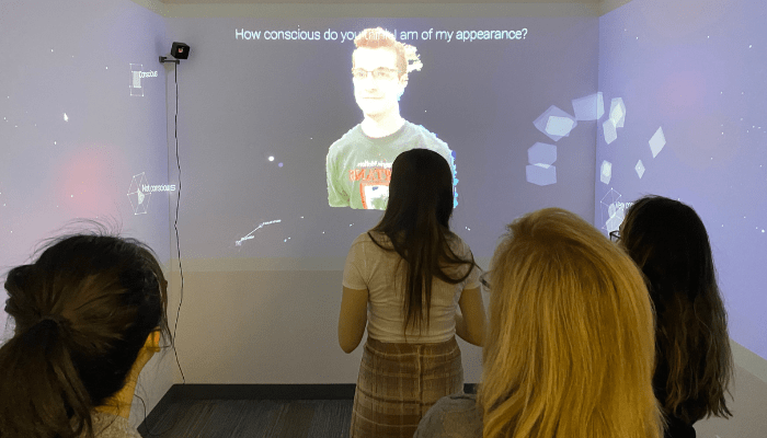 Visitors look towards a screen in a dark room. The screen shows an image of a man with the text "How conscious do you think I am of my appearance?"