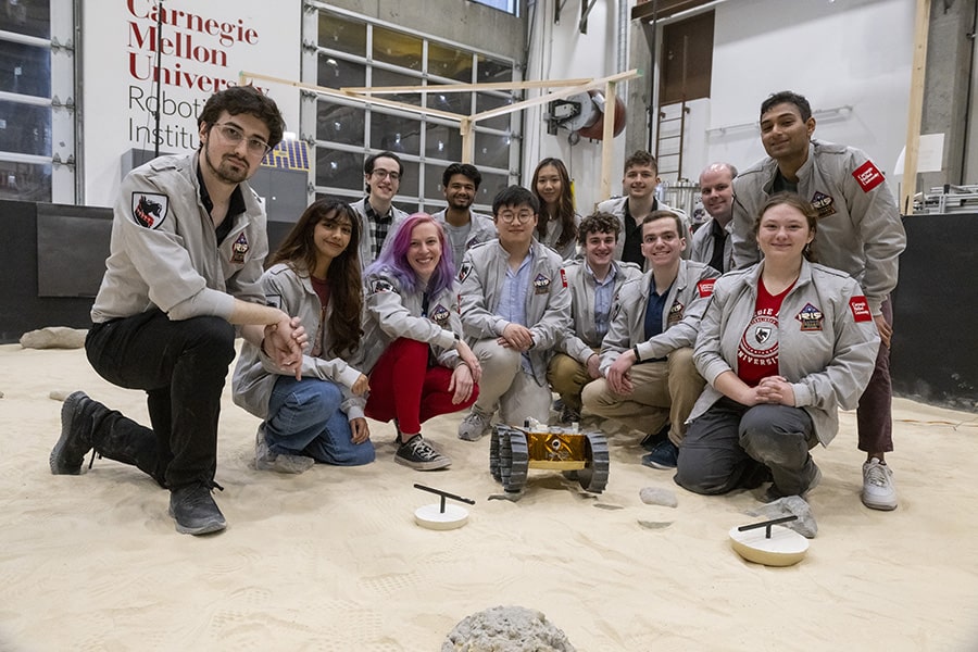 Students in gray IRIS/CMU branded jackets kneel in a lab. The floor is covered in sand and in the center of their group is a small lunar rover.